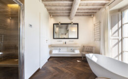 Modern bathroom featuring a freestanding bathtub, stylish vanity with a large mirror, and a walk-in shower. The space showcases elegant wooden flooring, exposed ceiling beams, and natural light streaming through the windows, creating a serene and luxurious atmosphere.