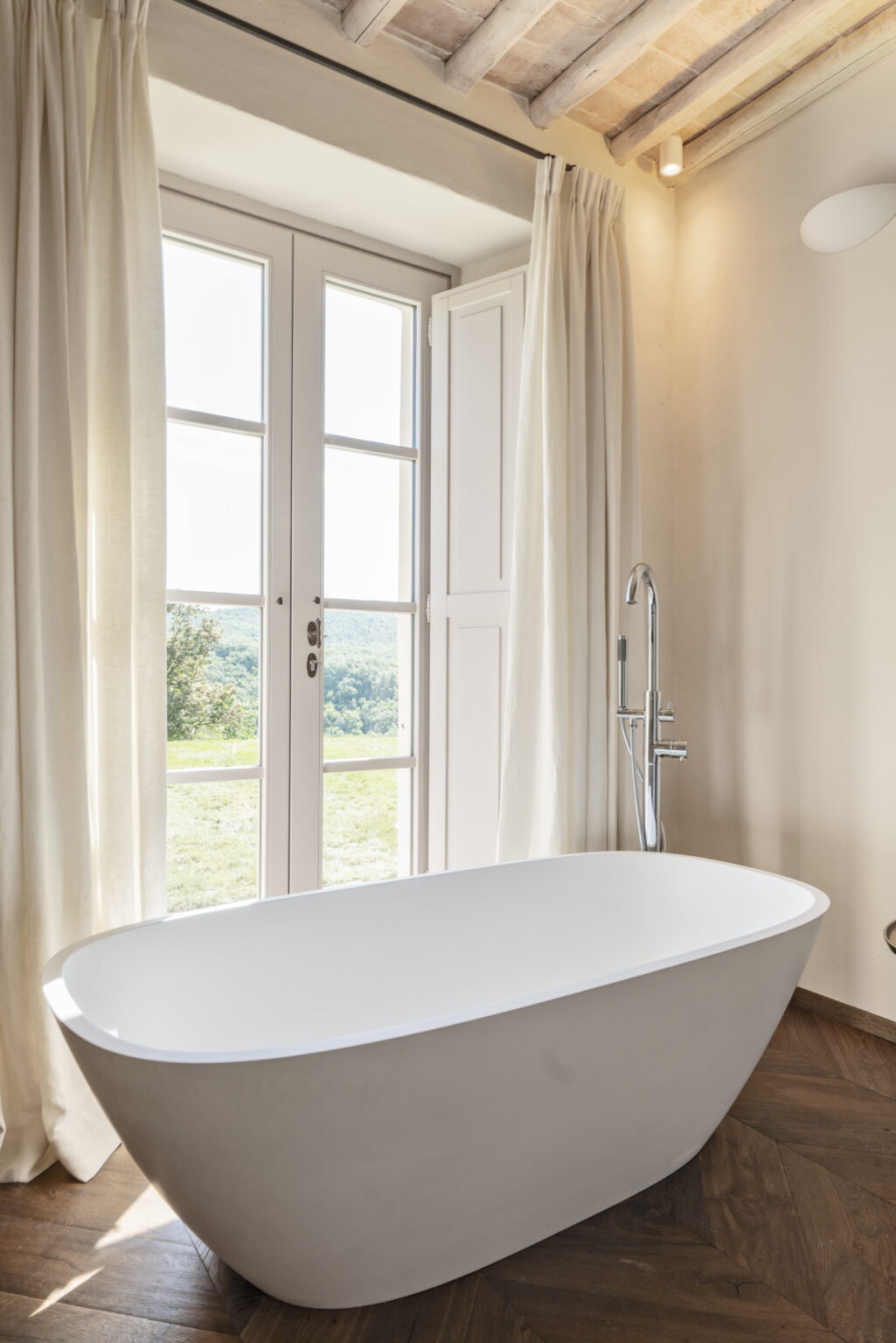 Modern bathroom featuring a freestanding white bathtub, elegant wooden flooring, and large windows with sheer curtains that offer a scenic view of the outdoors. The space is well-lit and exudes a serene, relaxing atmosphere. | Only on SopranoVillas