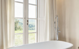 Modern bathroom featuring a freestanding white bathtub, elegant wooden flooring, and large windows with sheer curtains that offer a scenic view of the outdoors. The space is well-lit and exudes a serene, relaxing atmosphere.