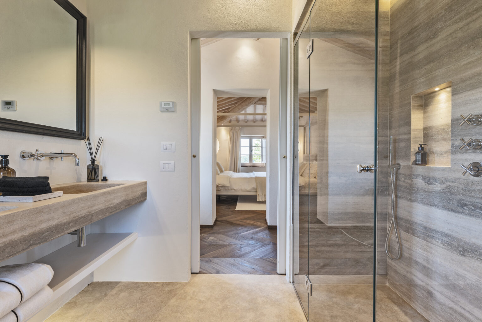 Modern bathroom interior featuring a stone sink, stylish mirror, and glass shower. The open doorway reveals a cozy bedroom with natural light and wooden flooring, enhancing the inviting atmosphere. Ideal for showcasing contemporary home design. | Only on SopranoVillas