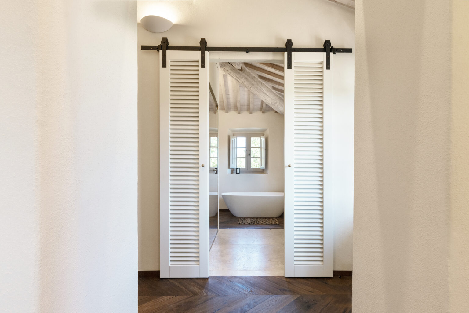 Interior view of a modern bathroom featuring a freestanding bathtub, framed by elegant white louvered sliding doors. The room showcases wooden beams and stylish flooring, creating a warm and inviting atmosphere. | Only on SopranoVillas