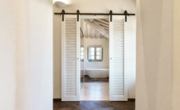Interior view of a modern bathroom featuring a freestanding bathtub, framed by elegant white louvered sliding doors. The room showcases wooden beams and stylish flooring, creating a warm and inviting atmosphere.