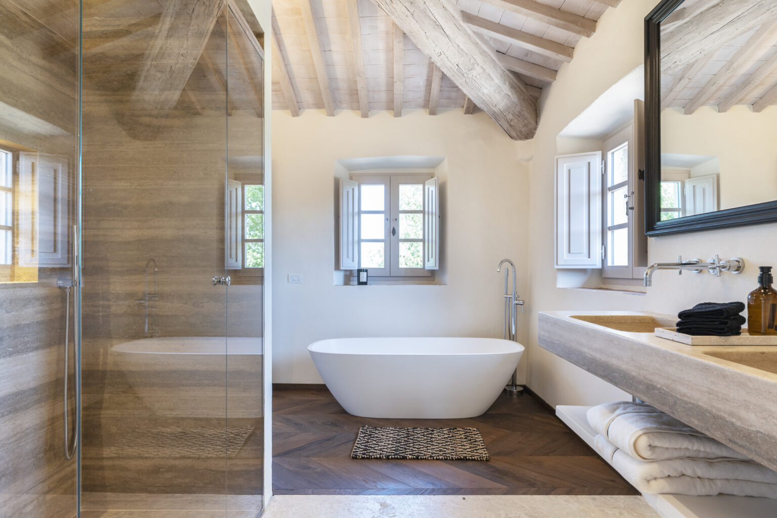 Modern bathroom interior featuring a freestanding white bathtub, a glass shower enclosure, and a double sink vanity with natural stone countertops. The room boasts wooden beam ceilings, stylish decor, and large windows allowing natural light to illuminate the space. Elegant towels and a decorative rug add to the aesthetic appeal. | Only on SopranoVillas