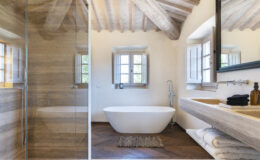 Modern bathroom interior featuring a freestanding white bathtub, a glass shower enclosure, and a double sink vanity with natural stone countertops. The room boasts wooden beam ceilings, stylish decor, and large windows allowing natural light to illuminate the space. Elegant towels and a decorative rug add to the aesthetic appeal.