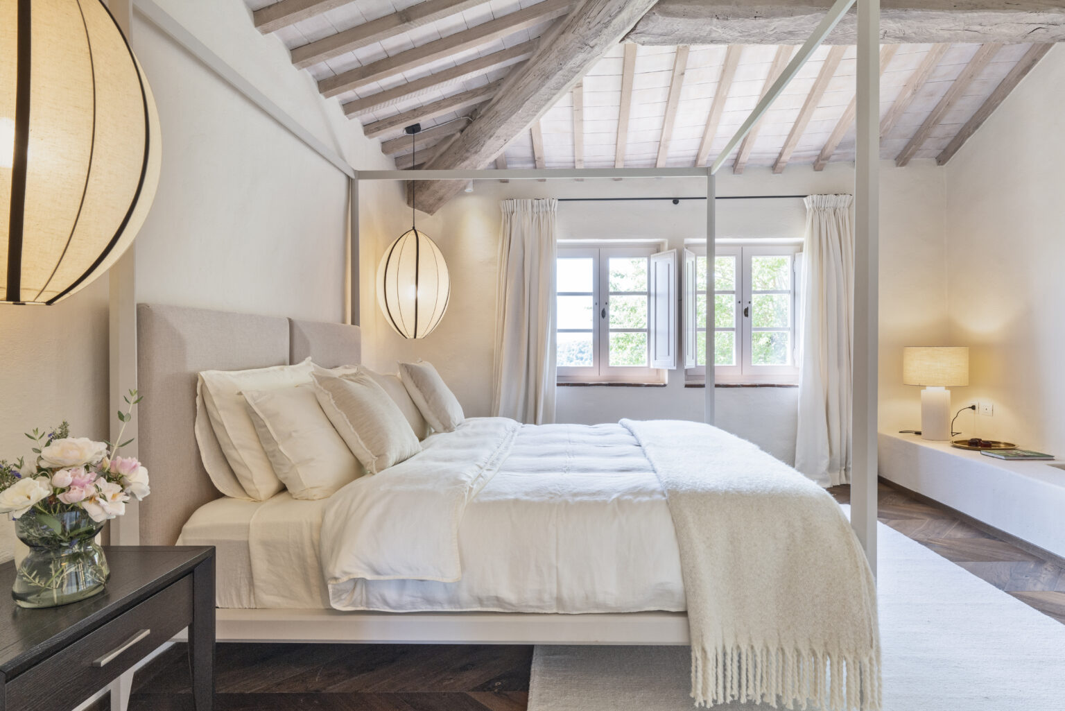 Modern bedroom interior featuring a stylish four-poster bed with soft white linens and decorative pillows. The room boasts high wooden ceilings, large windows with natural light, and elegant pendant lighting. A cozy bedside table holds a vase of flowers, complementing the serene atmosphere. | Only on SopranoVillas