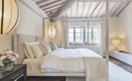 Modern bedroom interior featuring a stylish four-poster bed with soft white linens and decorative pillows. The room boasts high wooden ceilings, large windows with natural light, and elegant pendant lighting. A cozy bedside table holds a vase of flowers, complementing the serene atmosphere.
