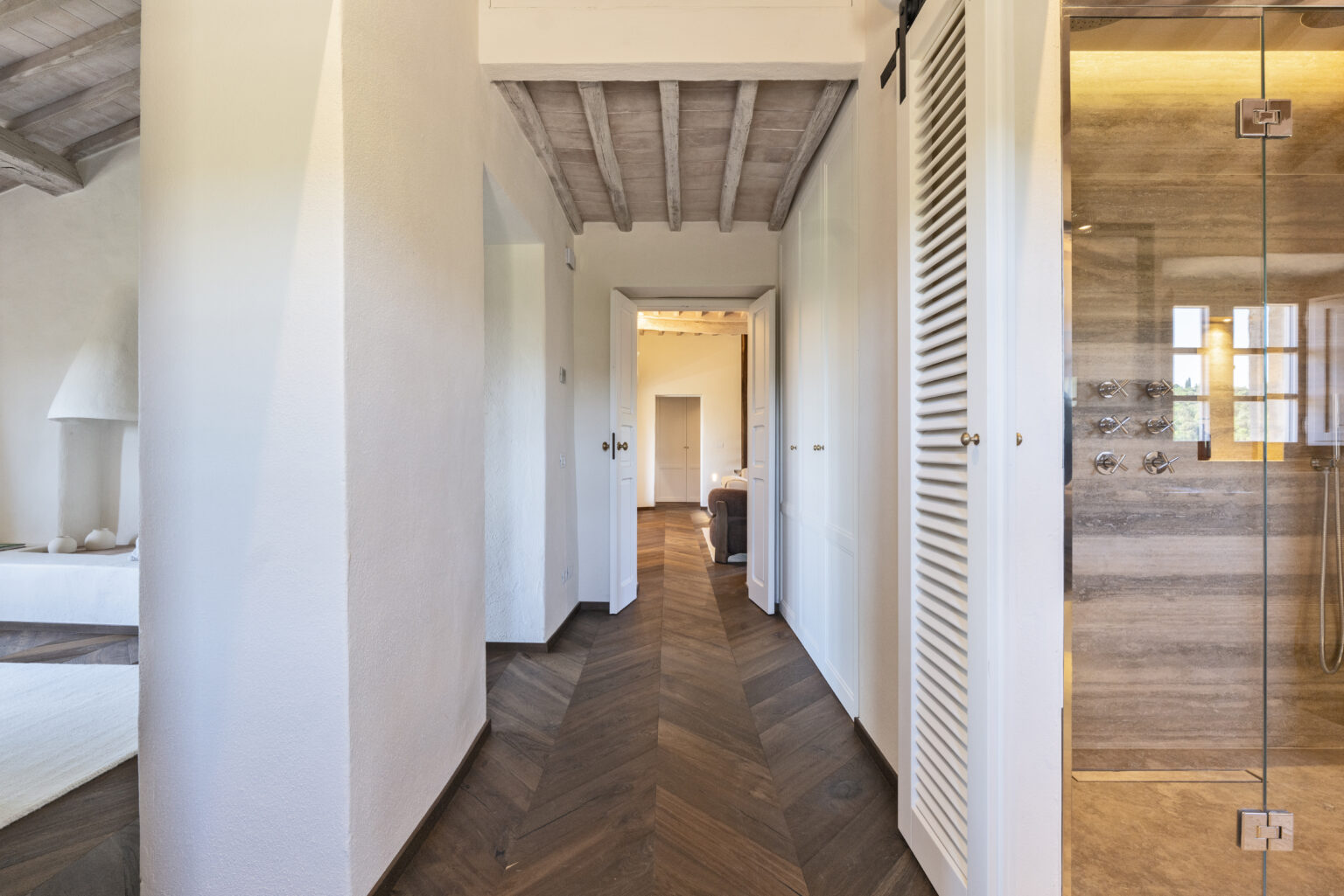 Modern interior hallway featuring a wooden herringbone floor, white walls, and a glass shower. The space is illuminated by natural light, showcasing a minimalist design with a cozy atmosphere. | Only on SopranoVillas