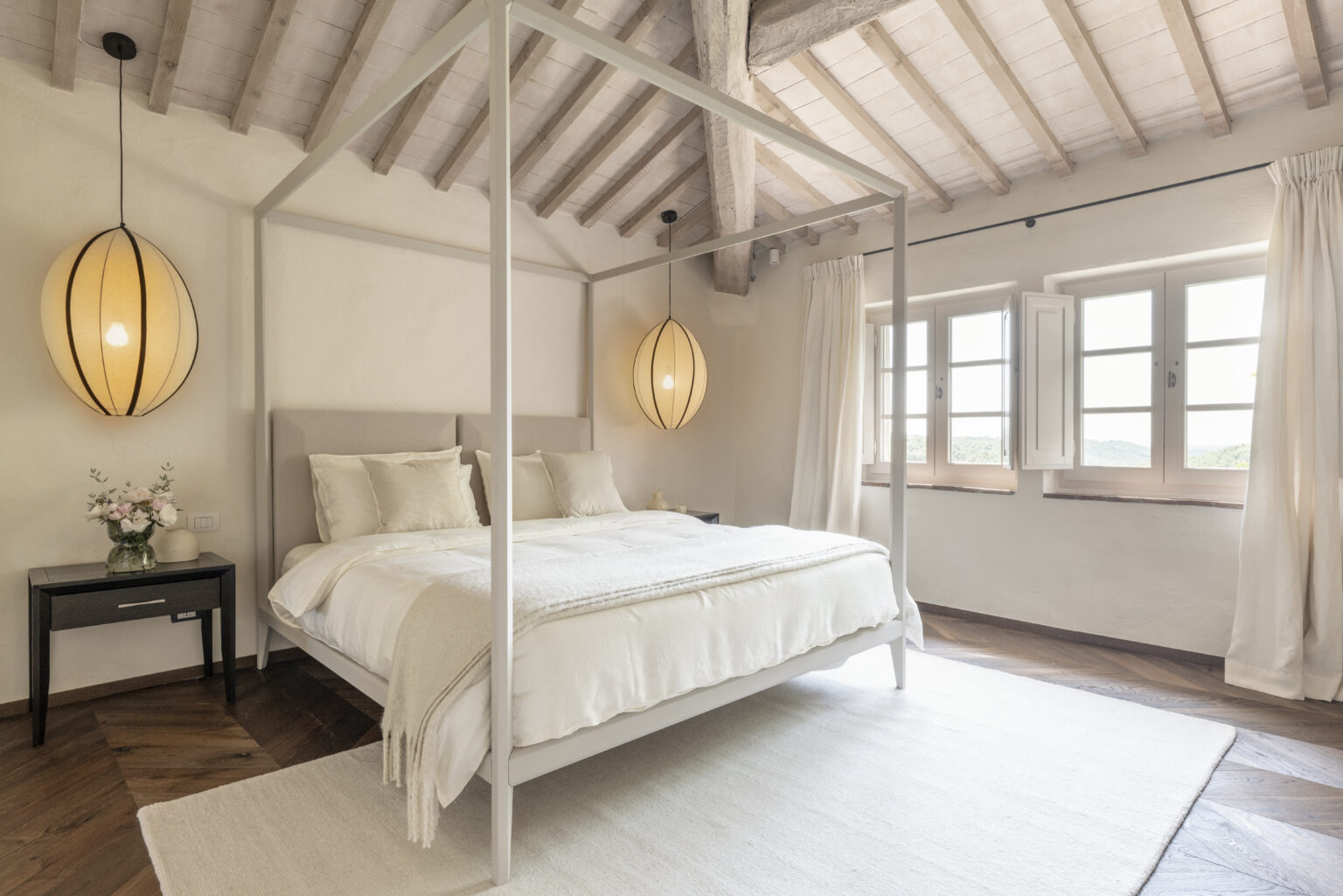 Modern bedroom featuring a four-poster bed with soft linens, a stylish pendant light, and large windows showcasing a scenic view. The space is decorated with neutral tones and natural wood accents, creating a serene and inviting atmosphere. | Only on SopranoVillas
