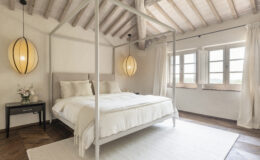 Modern bedroom featuring a four-poster bed with soft linens, a stylish pendant light, and large windows showcasing a scenic view. The space is decorated with neutral tones and natural wood accents, creating a serene and inviting atmosphere.