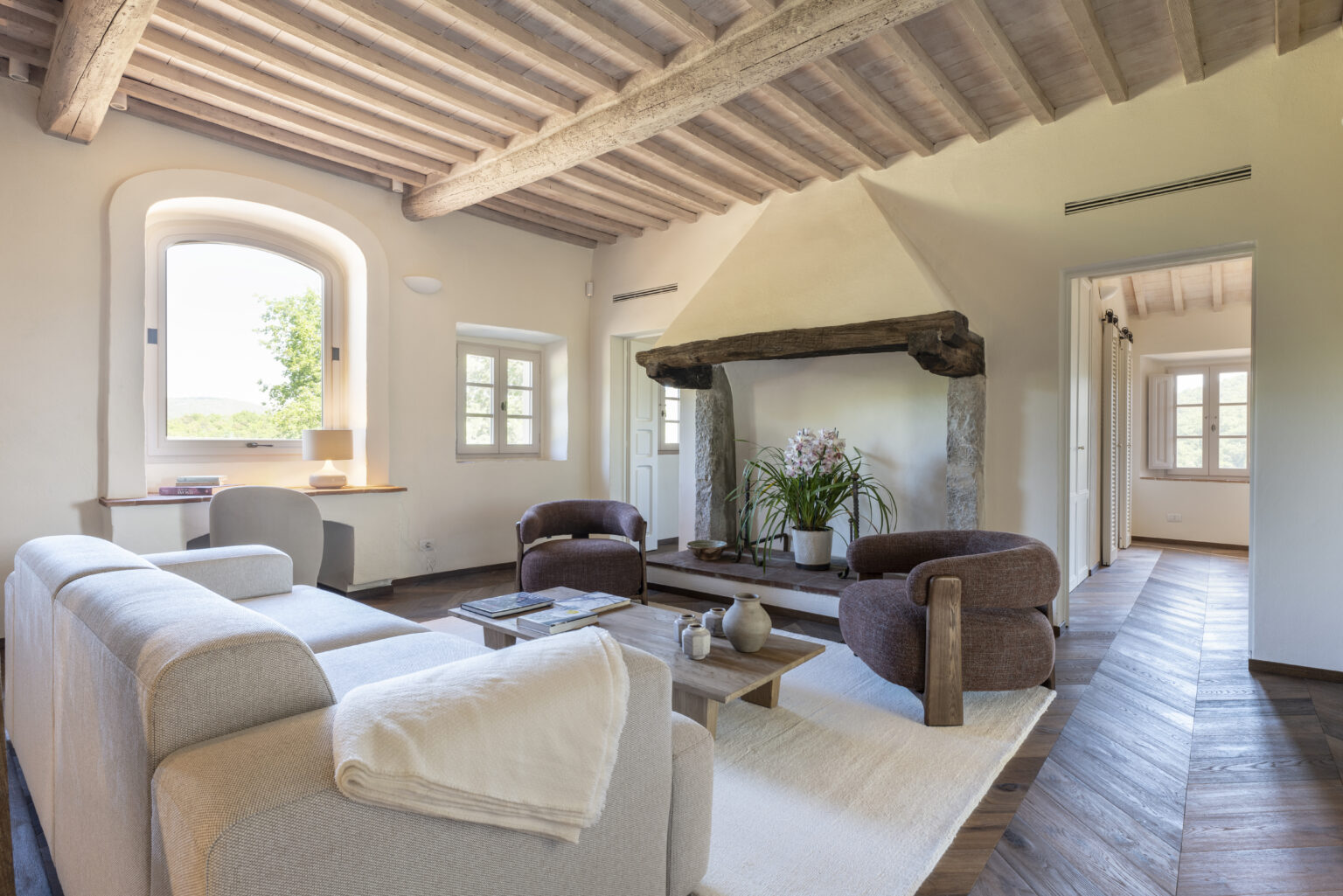 Spacious and inviting living room featuring a cozy seating area with a light-colored sofa and two brown armchairs, a wooden coffee table, and a stylish fireplace with a stone base. Large windows allow natural light to illuminate the room, complemented by rustic wooden beams on the ceiling and elegant decor, creating a warm and welcoming atmosphere. | Only on SopranoVillas