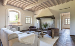 Spacious and inviting living room featuring a cozy seating area with a light-colored sofa and two brown armchairs, a wooden coffee table, and a stylish fireplace with a stone base. Large windows allow natural light to illuminate the room, complemented by rustic wooden beams on the ceiling and elegant decor, creating a warm and welcoming atmosphere.