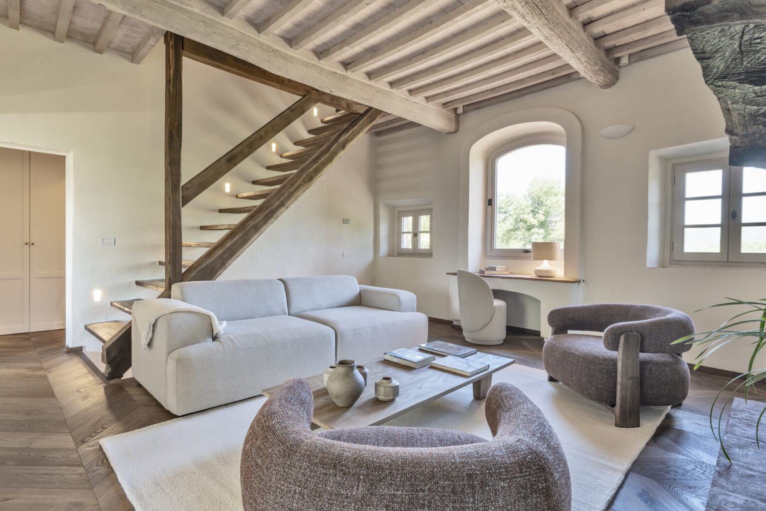 Modern living room interior featuring a cozy sofa, stylish armchairs, and a wooden coffee table. A wooden staircase leads to an upper level, while large windows provide natural light and a view of greenery outside. The space combines contemporary design with rustic elements, creating a warm and inviting atmosphere. | Only on SopranoVillas