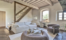 Modern living room interior featuring a cozy sofa, stylish armchairs, and a wooden coffee table. A wooden staircase leads to an upper level, while large windows provide natural light and a view of greenery outside. The space combines contemporary design with rustic elements, creating a warm and inviting atmosphere.