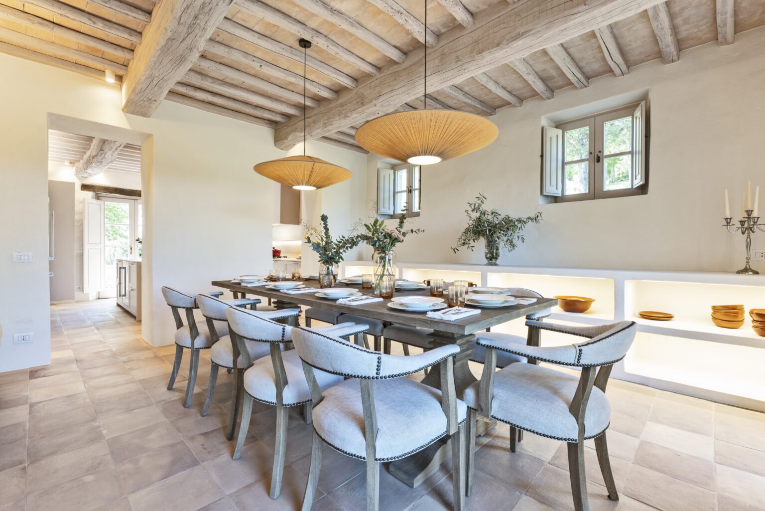 Modern dining room featuring a rustic wooden table surrounded by upholstered chairs, illuminated by stylish pendant lights. The space includes natural decor elements, such as greenery in vases and a minimalist side shelf displaying tableware, complemented by warm, neutral tones and exposed wooden beams in the ceiling. | Only on SopranoVillas