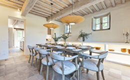 Modern dining room featuring a rustic wooden table surrounded by upholstered chairs, illuminated by stylish pendant lights. The space includes natural decor elements, such as greenery in vases and a minimalist side shelf displaying tableware, complemented by warm, neutral tones and exposed wooden beams in the ceiling.
