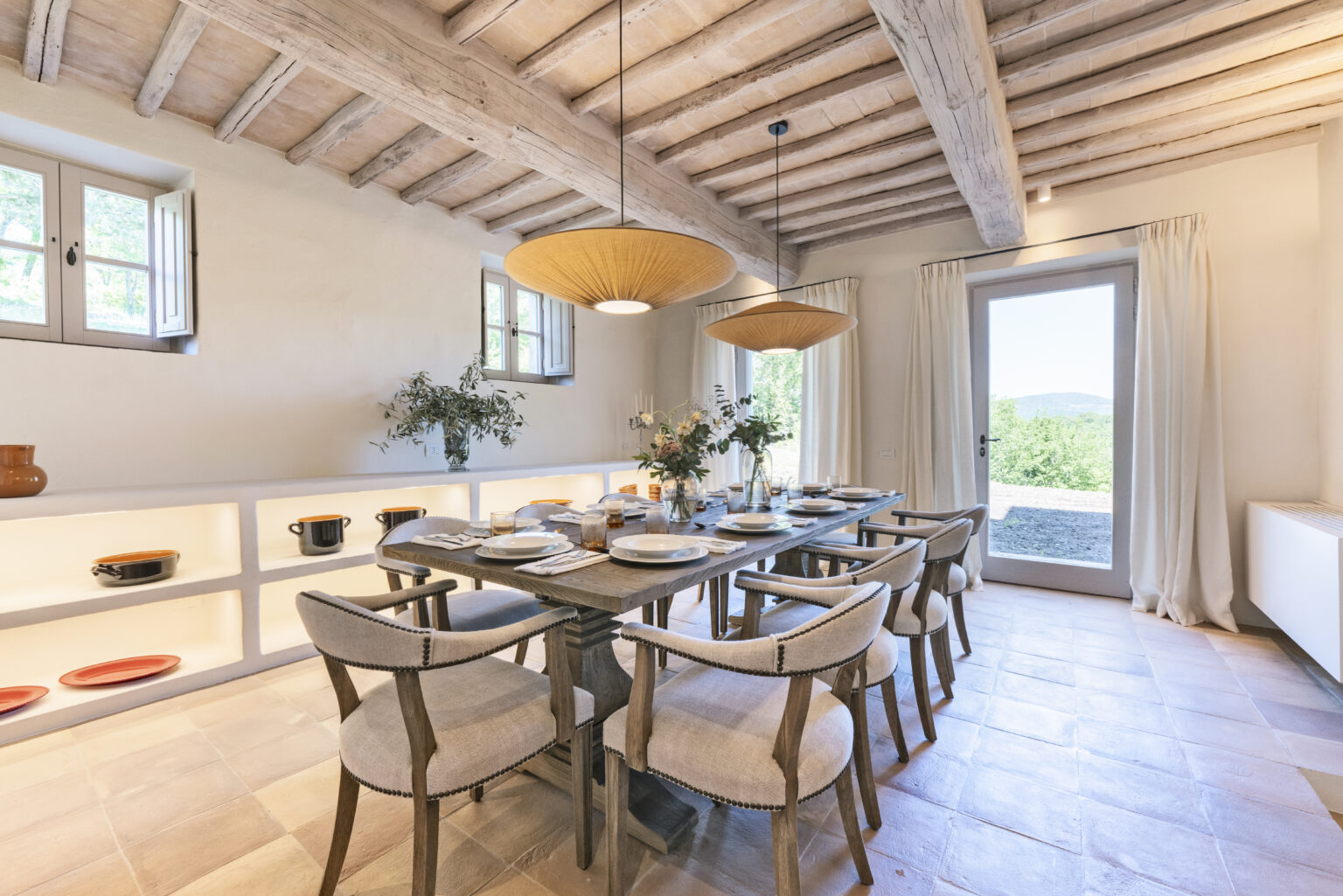 Modern dining room featuring a wooden table set for a meal, surrounded by stylish chairs. Natural light floods in through large windows, highlighting the rustic wooden ceiling and elegant decor, including a vase of flowers and shelves displaying ceramic dishes. | Only on SopranoVillas