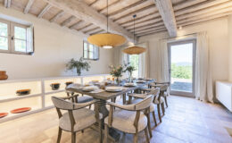 Modern dining room featuring a wooden table set for a meal, surrounded by stylish chairs. Natural light floods in through large windows, highlighting the rustic wooden ceiling and elegant decor, including a vase of flowers and shelves displaying ceramic dishes.