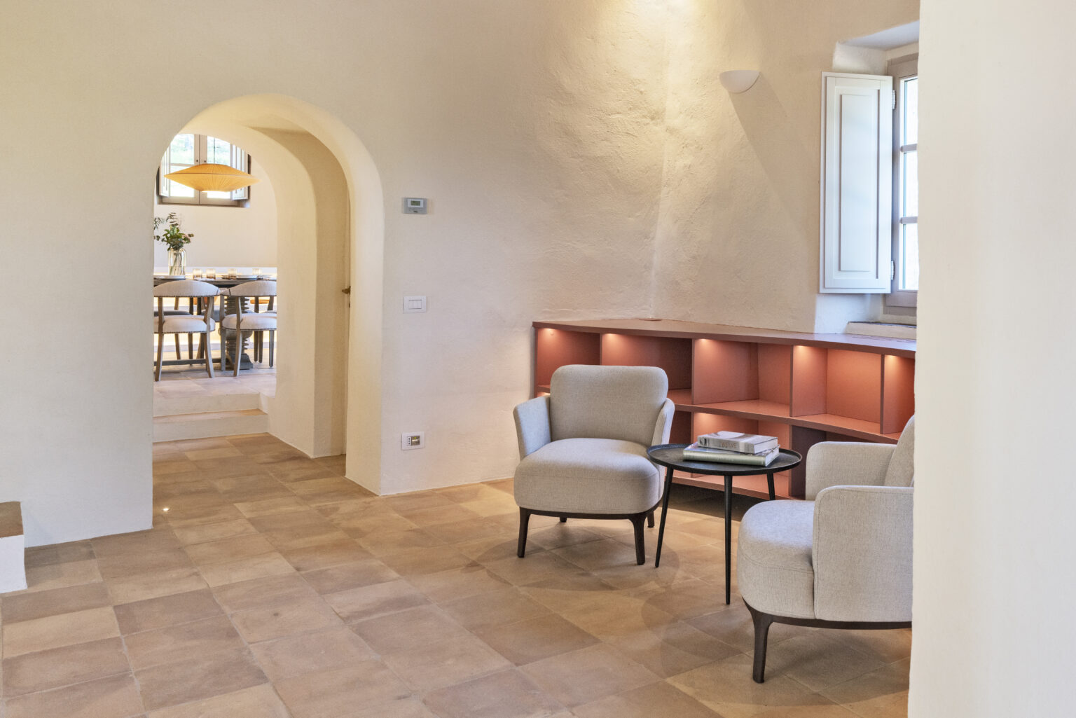 Bright and airy interior of a modern living space featuring two gray armchairs, a small round table with books, and a soft color palette. An arched doorway leads to a dining area with a wooden table and elegant lighting, enhancing the open and inviting atmosphere. | Only on SopranoVillas