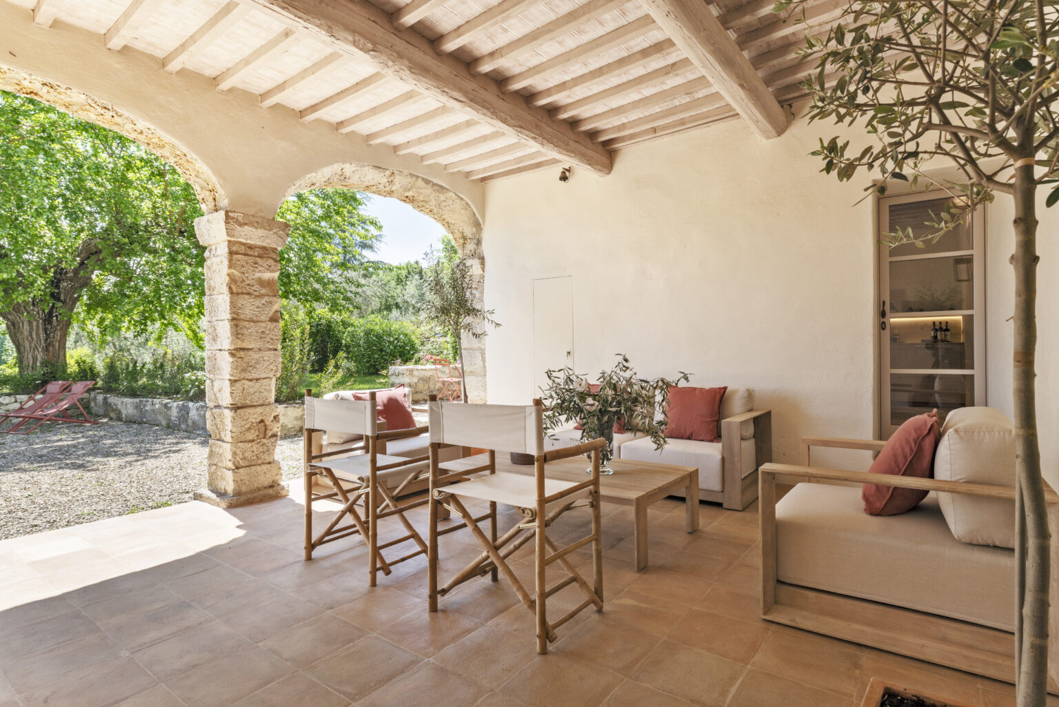 Cozy outdoor seating area featuring wooden chairs and a sofa with cushions, surrounded by lush greenery and stone columns, perfect for relaxation in a tranquil garden setting. | Only on SopranoVillas