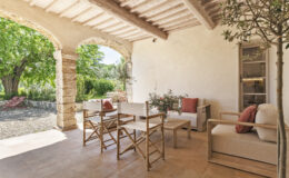 Cozy outdoor seating area featuring wooden chairs and a sofa with cushions, surrounded by lush greenery and stone columns, perfect for relaxation in a tranquil garden setting.