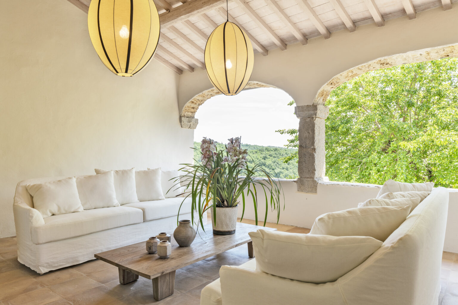 Bright and airy living room with white sofas, a wooden coffee table, and decorative plants, featuring large windows that open to a scenic view of greenery and a rustic ceiling with pendant lighting. Ideal for relaxation and modern home decor inspiration. | Only on SopranoVillas