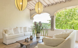 Bright and airy living room with white sofas, a wooden coffee table, and decorative plants, featuring large windows that open to a scenic view of greenery and a rustic ceiling with pendant lighting. Ideal for relaxation and modern home decor inspiration.