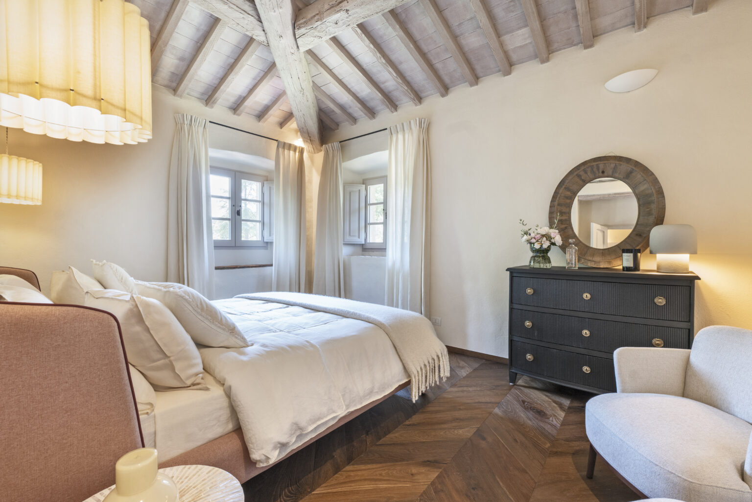 Cozy bedroom interior featuring a comfortable bed with plush pillows, a stylish black dresser with decorative mirror, and elegant lighting. The room has wooden beams, large windows with sheer curtains, and warm hardwood flooring, creating a serene and inviting atmosphere. Ideal for relaxation and restful sleep. | Only on SopranoVillas