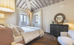 Cozy bedroom interior featuring a comfortable bed with plush pillows, a stylish black dresser with decorative mirror, and elegant lighting. The room has wooden beams, large windows with sheer curtains, and warm hardwood flooring, creating a serene and inviting atmosphere. Ideal for relaxation and restful sleep.