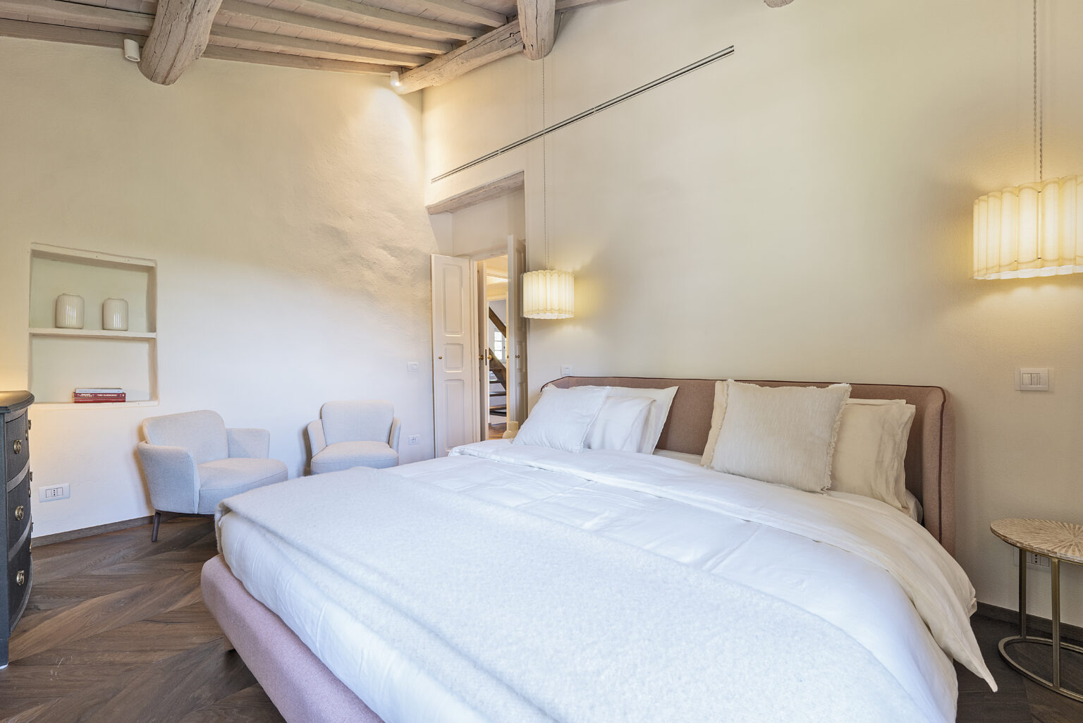 Modern and cozy bedroom featuring a plush bed with white bedding, two elegant armchairs, and stylish lighting. The room showcases wooden beams and a warm color palette, creating a serene atmosphere. Ideal for relaxation and comfort in a contemporary setting. | Only on SopranoVillas