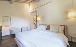 Modern and cozy bedroom featuring a plush bed with white bedding, two elegant armchairs, and stylish lighting. The room showcases wooden beams and a warm color palette, creating a serene atmosphere. Ideal for relaxation and comfort in a contemporary setting.