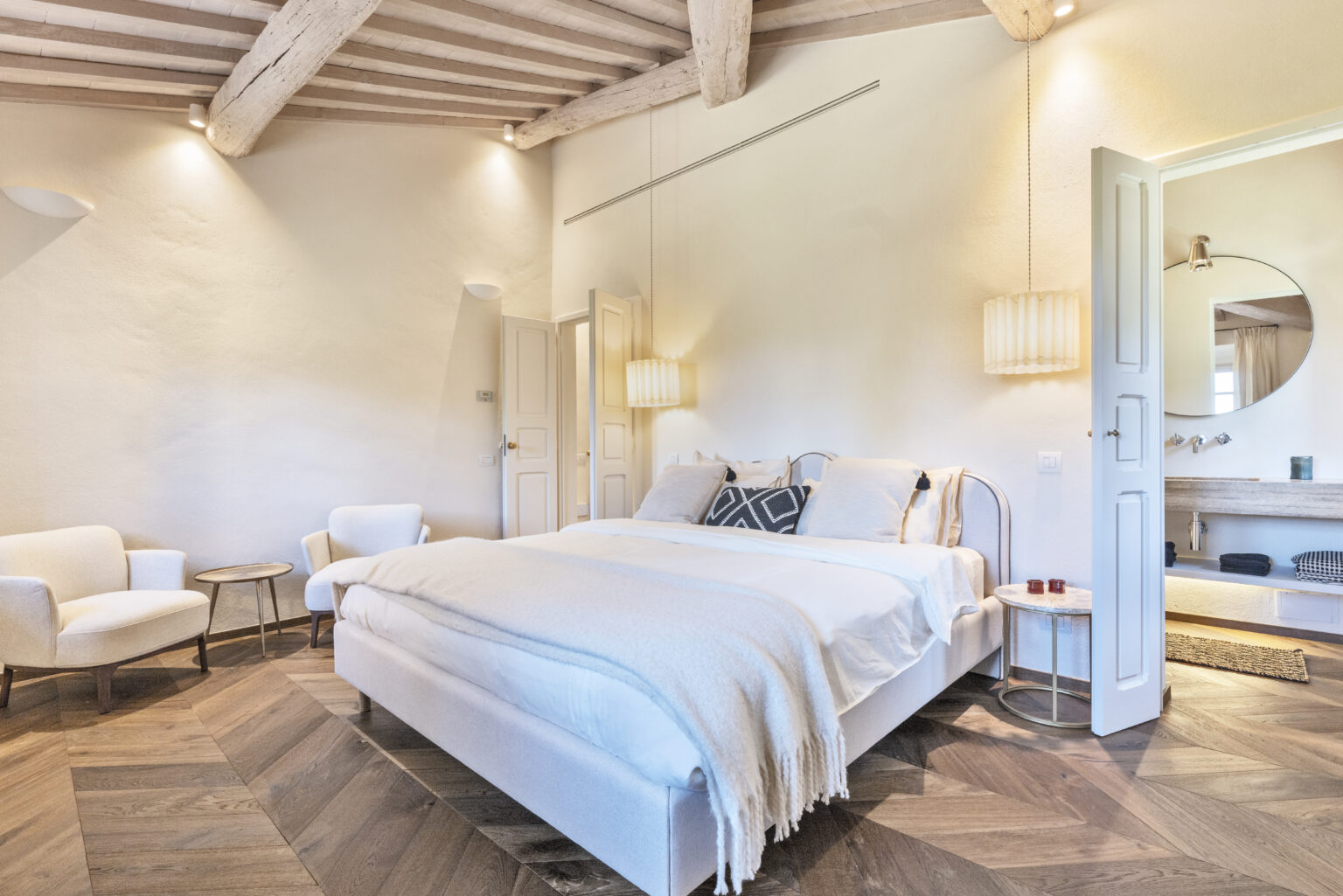 Modern bedroom interior featuring a plush bed with white linens, decorative pillows, and a cozy throw. The room has wooden beams, stylish lighting, and comfortable seating arrangements, creating a serene and inviting atmosphere. A doorway leads to a bathroom area with a round mirror and wooden vanity. | Only on SopranoVillas