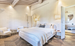 Modern bedroom interior featuring a plush bed with white linens, decorative pillows, and a cozy throw. The room has wooden beams, stylish lighting, and comfortable seating arrangements, creating a serene and inviting atmosphere. A doorway leads to a bathroom area with a round mirror and wooden vanity.