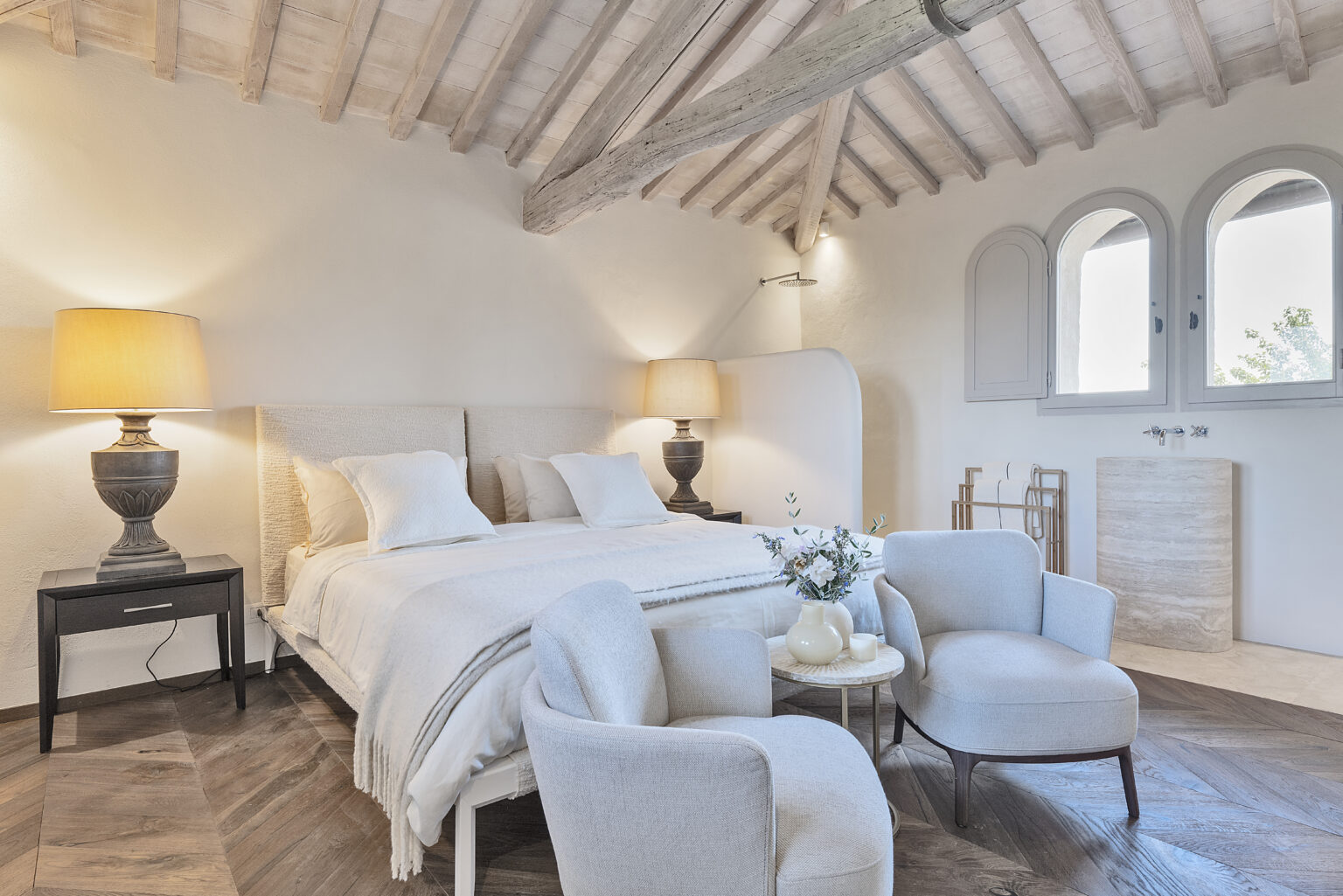 Modern bedroom interior featuring a cozy bed with textured headboard, stylish bedside lamp, and elegant seating area. The room showcases wooden beams, natural light from arched windows, and chic decor elements, creating a serene and inviting atmosphere. | Only on SopranoVillas
