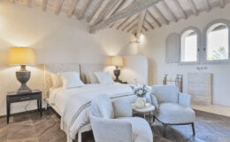 Modern bedroom interior featuring a cozy bed with textured headboard, stylish bedside lamp, and elegant seating area. The room showcases wooden beams, natural light from arched windows, and chic decor elements, creating a serene and inviting atmosphere.