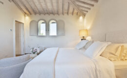 Cozy and elegant bedroom featuring a spacious bed with white linens, soft pillows, and a light throw. The room has a rustic wooden ceiling, tasteful lamp, and decorative flower arrangement on a side table. Bright natural light filters through arched windows, enhancing the serene atmosphere.
