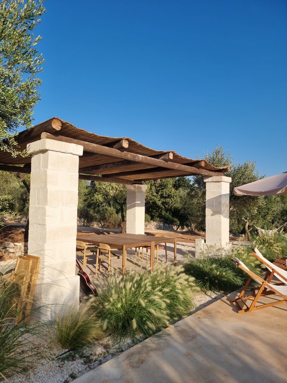 Outdoor dining area with a wooden pergola and stone columns, surrounded by lush greenery and a clear blue sky, ideal for gatherings and relaxation. | Only on SopranoVillas