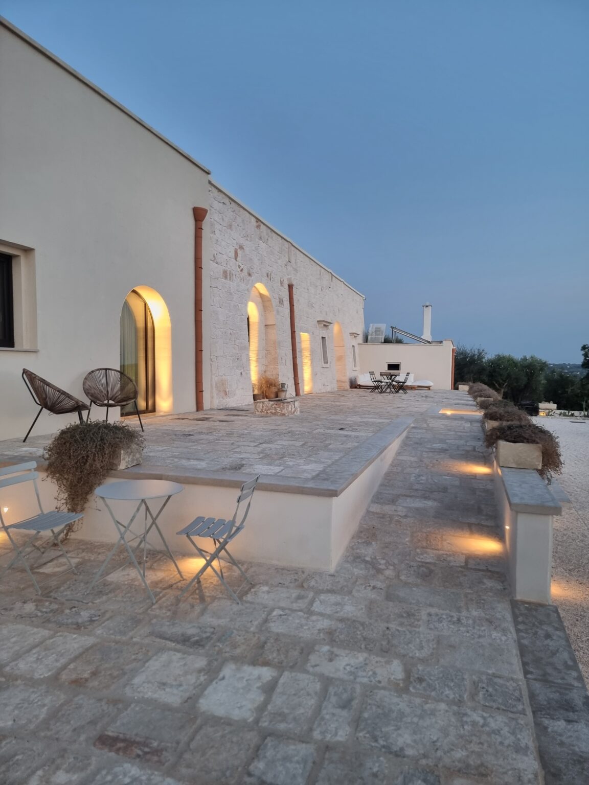 Modern stone villa exterior at dusk, featuring arched windows and a spacious patio with stylish seating. Soft lighting highlights the stone pathway, creating an inviting atmosphere for outdoor gatherings. Lush greenery surrounds the property, enhancing its tranquil ambiance. | Only on SopranoVillas