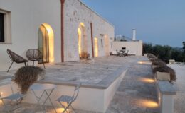Modern stone villa exterior at dusk, featuring arched windows and a spacious patio with stylish seating. Soft lighting highlights the stone pathway, creating an inviting atmosphere for outdoor gatherings. Lush greenery surrounds the property, enhancing its tranquil ambiance.
