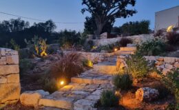 Stone pathway illuminated by warm lights, leading through a beautifully landscaped garden at dusk, featuring lush plants and a large tree in the background.