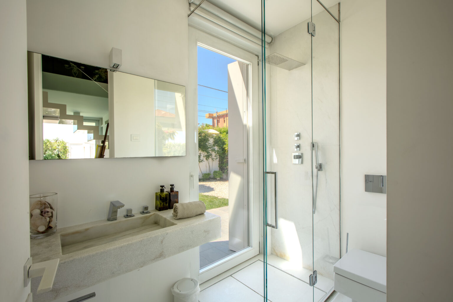 Modern bathroom design featuring a minimalist aesthetic with a marble sink, large mirror, and glass shower enclosure. Natural light floods the space through a door leading to an outdoor garden. Stylish toiletries are neatly arranged on the countertop, enhancing the contemporary feel. | Only on SopranoVillas