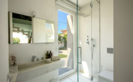 Modern bathroom design featuring a minimalist aesthetic with a marble sink, large mirror, and glass shower enclosure. Natural light floods the space through a door leading to an outdoor garden. Stylish toiletries are neatly arranged on the countertop, enhancing the contemporary feel.