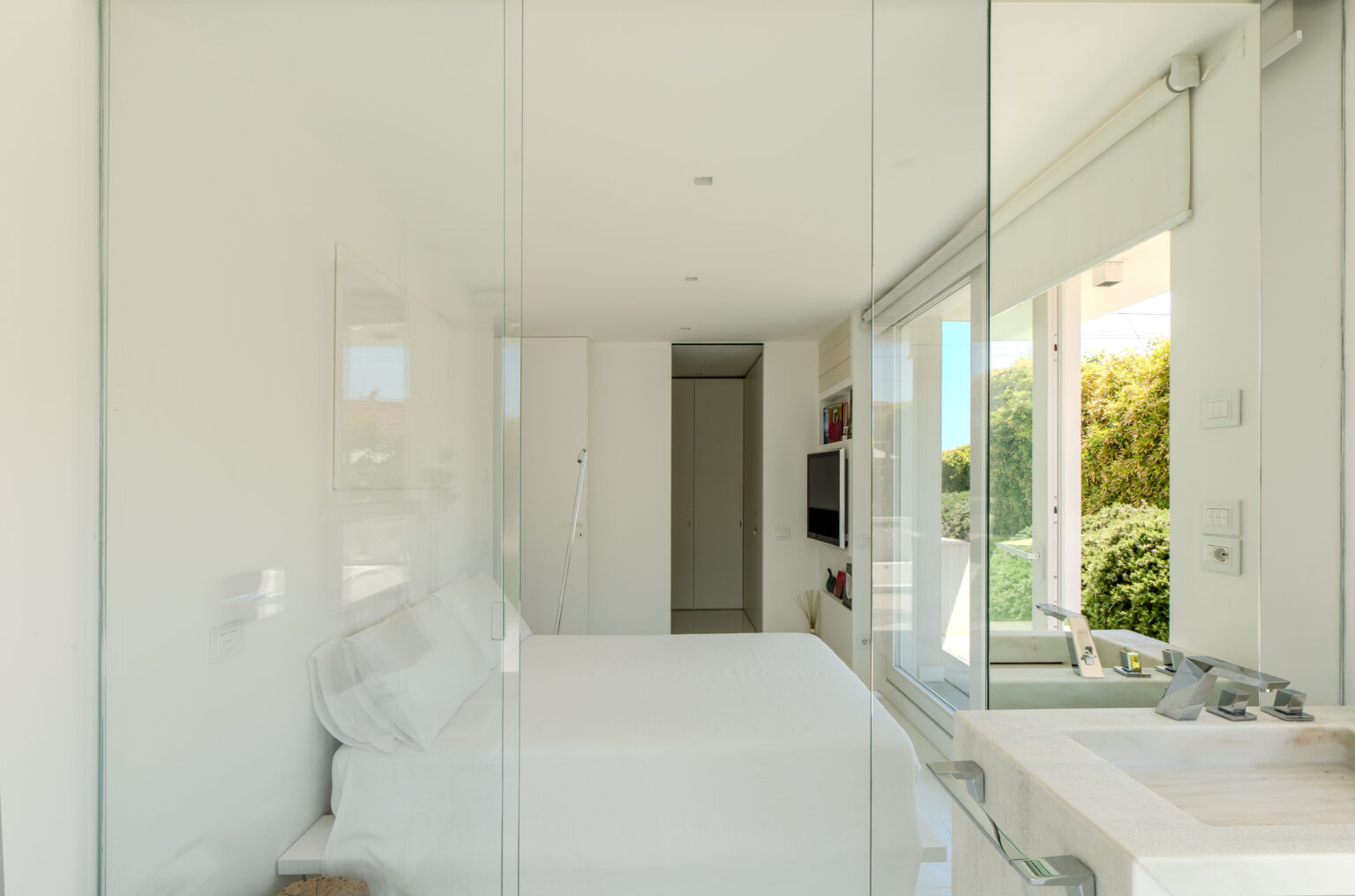 Modern bedroom with glass walls, featuring a minimalist design, white bedding, and natural light streaming in from large windows. The space includes a compact television and stylish decorative elements, creating a serene atmosphere. | Only on SopranoVillas