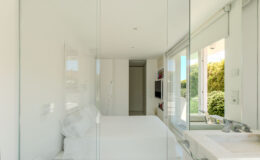Modern bedroom with glass walls, featuring a minimalist design, white bedding, and natural light streaming in from large windows. The space includes a compact television and stylish decorative elements, creating a serene atmosphere.