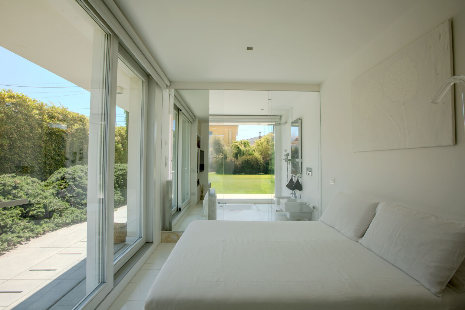 Modern bedroom with large glass windows, featuring a white bed and minimalist decor, overlooking a green garden. Natural light fills the space, creating a serene atmosphere. | Only on SopranoVillas