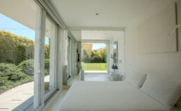 Modern bedroom with large glass windows, featuring a white bed and minimalist decor, overlooking a green garden. Natural light fills the space, creating a serene atmosphere.