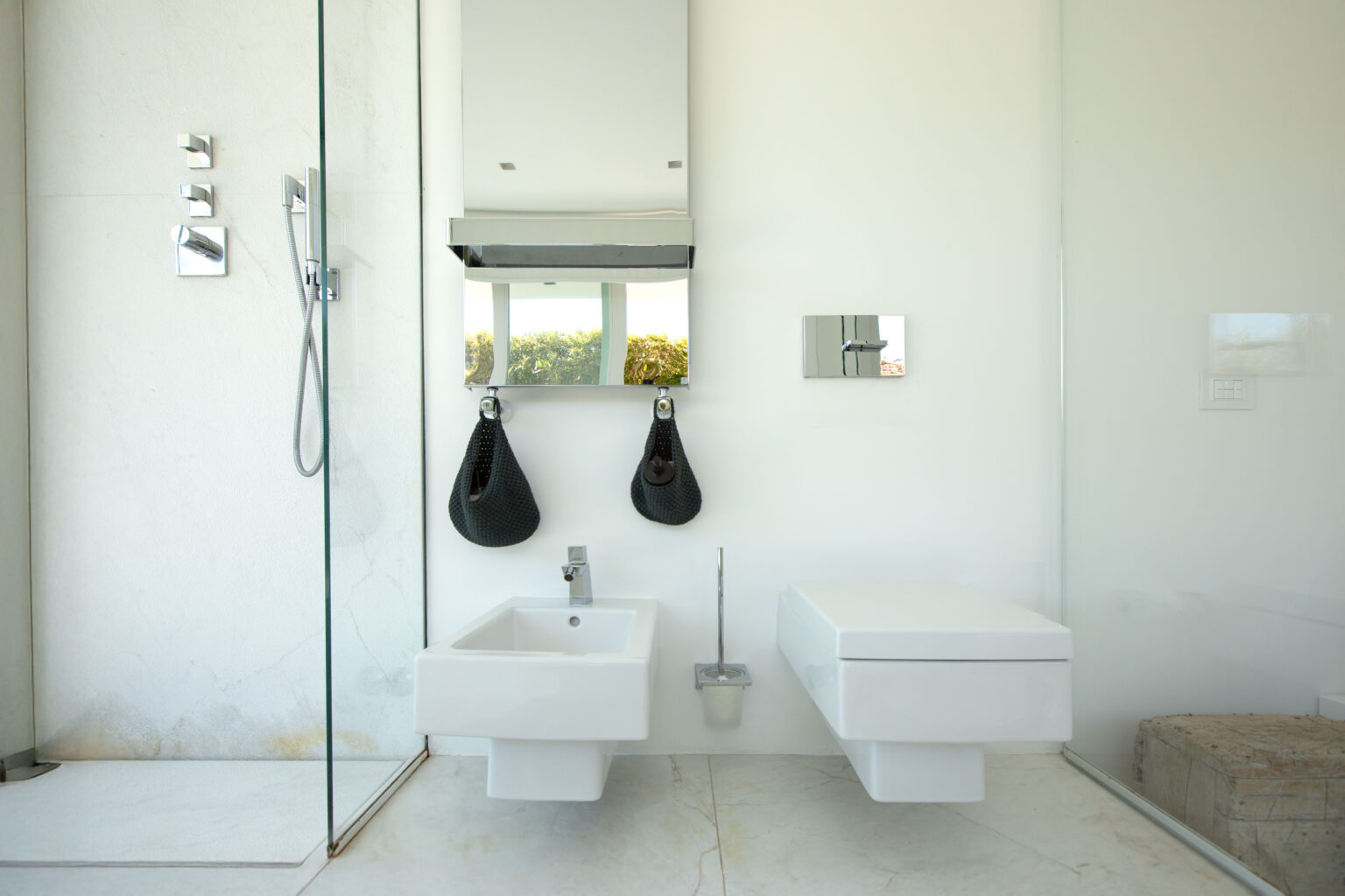 Modern bathroom interior featuring a glass shower, wall-mounted toilet, and sleek sink. The design emphasizes minimalism with white walls, natural stone flooring, and stylish fixtures, complemented by decorative hanging bags and a large mirror. | Only on SopranoVillas