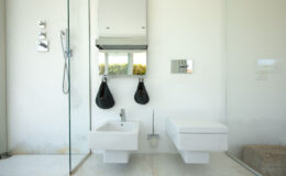 Modern bathroom interior featuring a glass shower, wall-mounted toilet, and sleek sink. The design emphasizes minimalism with white walls, natural stone flooring, and stylish fixtures, complemented by decorative hanging bags and a large mirror.