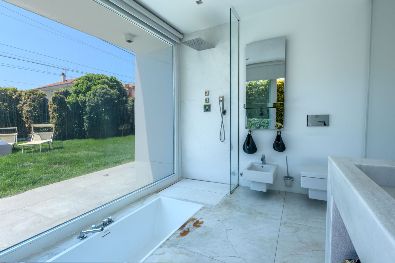 Modern bathroom interior featuring a sleek design with a glass shower, minimalist sink, and a large window overlooking a green garden. The space includes a bathtub and contemporary fixtures, emphasizing a bright and airy atmosphere. | Only on SopranoVillas