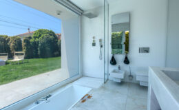 Modern bathroom interior featuring a sleek design with a glass shower, minimalist sink, and a large window overlooking a green garden. The space includes a bathtub and contemporary fixtures, emphasizing a bright and airy atmosphere.