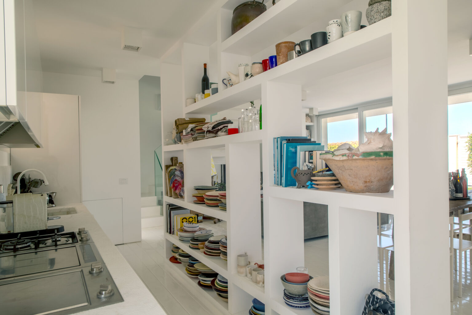 Modern kitchen interior featuring a white shelving unit filled with colorful plates, mugs, and glassware, alongside neatly stacked cookbooks and decorative items. The minimalist design enhances the spacious feel of the kitchen, with natural light streaming through large windows. | Only on SopranoVillas